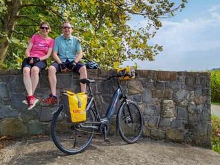 Two cyclists in sunglasses sitting on a stone wall with their bicycle parked in front, on the Alpe Adria cycle route near Grado.