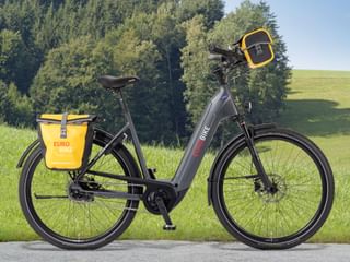 Dunkelgraues Eurobike Leih-E-Bike mit gelben Taschen am Gepäckträger und Lenker, am Straßenrand mit grüner Wiese und Wald im Hintergrund.