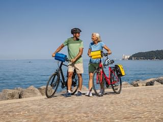 Couple with touring bikes on paved waterfront path near Trieste. Miramare Castle visible on distant hillside across calm blue Adriatic Sea.