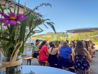 Employees seated at beer garden tables at an outdoor summer party. Pink flowers in foreground, rolling hills and blue sky in background.
