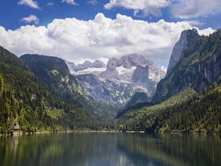 Lake Gosausee in Salzkammergut with forested mountains and Dachstein massif with snow-capped peaks under dramatic clouds.