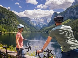 Two cyclists with helmets pause at Gosausee lake shore, viewing the Dachstein mountain range with snow-capped peaks and forested slopes.
