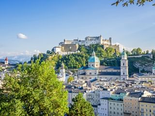 Salzburger Stadtbild mit historischen Gebäuden, barocken Kirchenkuppeln und der Festung Hohensalzburg auf einem Hügel. Grüne Bäume rahmen die Aussicht.