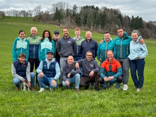 Group photo of 16 people in outdoor clothing on a green meadow. Rolling hills and forest visible in the background under cloudy sky.