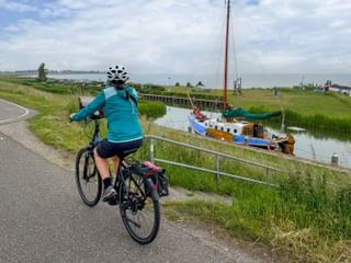 Female cyclist in turquoise jacket riding on paved dike path along IJsselmeer with traditional sailboat moored in canal and green marshland.
