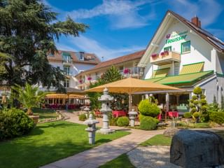 Gartenhotel Heusser in Bad Dürkheim mit weißer Fassade, grünen Markisen, Terrasse mit gelben Sonnenschirmen und Garten mit asiatischen Steinlaternen.