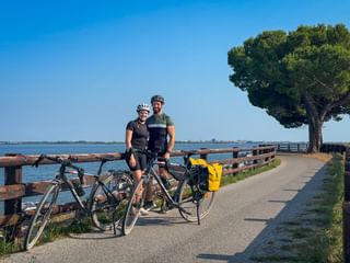 Two cyclists with helmets and touring bikes on coastal path with wooden fence, lagoon, and pine tree near Grado on the Alpe Adria cycle route.