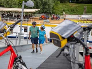 Zwei Radfahrer auf der Boarding-Rampe eines Donau-Flussschiffs in Melk. Fahrräder im Vordergrund, gelbes Schiffsgeländer mit 'Rad & Schiff'-Schriftzug.
