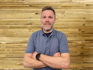 Stefan Krautgartner mit verschränkten Armen in grauem Poloshirt und Uhr, lächelnd vor einer Holzwand.