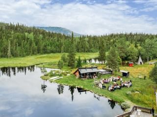 Luftaufnahme einer Versammlung am ruhigen See in Hemsedal Kommune. Menschen sitzen um eine Holzhütte, umgeben von grünem Wald und Bergen.