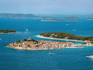 Aerial view of Primošten peninsula with terracotta roofs, church tower, and turquoise Adriatic Sea. Islands and mountains visible in background.