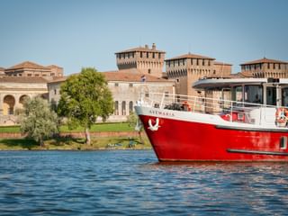 Rot-weißes Passagierschiff MS Ave Maria auf blauem Wasser mit historischen Festungsmauern und Türmen in Mantua, Italien unter klarem Himmel.