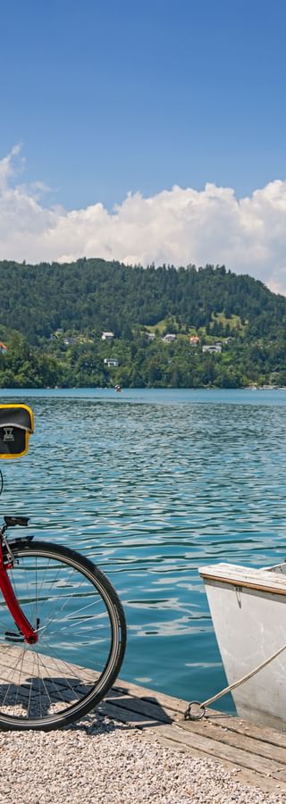 Two cyclists with helmets and bikes stand by Lake Bled near a boat. A church on an island and forested hills are visible in the background.