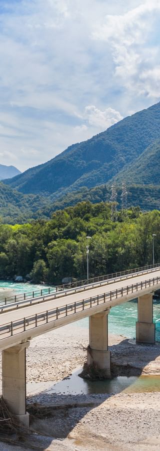 Radfahrer überqueren eine Betonbrücke über den türkisfarbenen Tagliamento bei Venzone, mit bewaldeten Bergen und einem kleinen Dorf im Hintergrund.