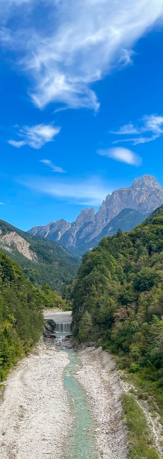 Alpental mit türkisfarbenem Fluss durch steiniges Flussbett, umgeben von bewaldeten Bergen unter blauem Himmel mit weißen Wolken.