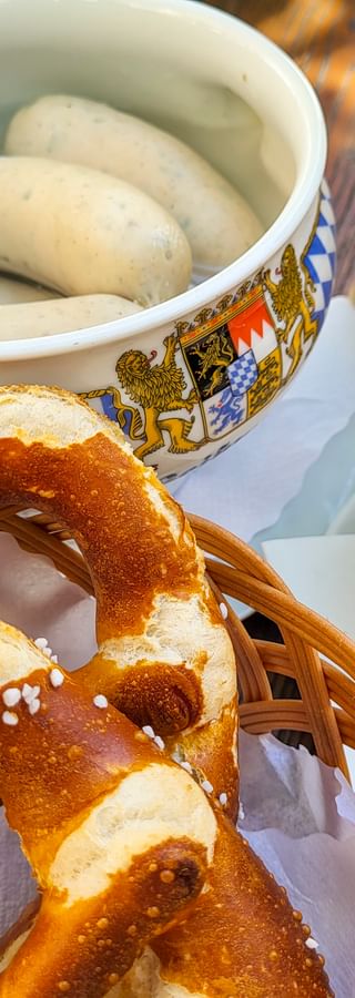 Traditionelles bayerisches Essen mit Weißwürsten in Keramikschale, Brezel mit Salz, Senf und Kellerbier auf Holztisch am Kochelsee.