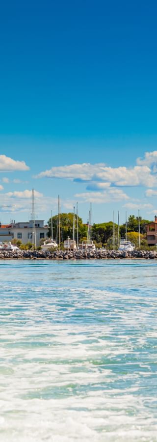 View of Grado's colorful waterfront buildings and marina from a ship, with turquoise water in foreground and blue sky with white clouds above.