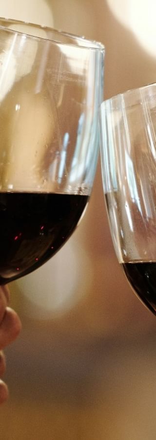 Zwei Hände halten Weingläser mit Rotwein und stoßen vor einem warmen, unscharfen Hintergrund in der Toskana an.