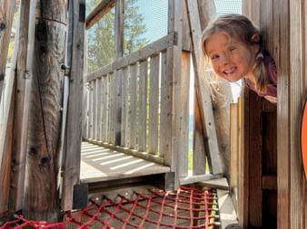 Lächelndes Mädchen schaut durch Holzspielplatz mit Kletternetz. Holzbrücke und Schutznetz im Hintergrund sichtbar.