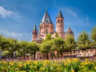 Mainzer Dom mit roten Sandsteintürmen über grünen Bäumen und buntem Blumengarten mit gelben und violetten Blüten unter blauem Himmel.