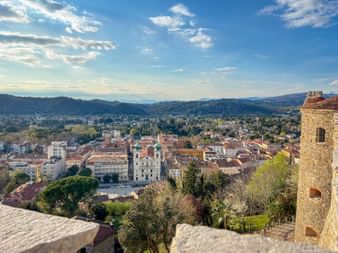 Aerial view of Gorizia with historic buildings, a white church, and stone tower in foreground. Rolling hills and blue sky in background.