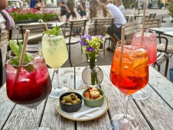 Four colorful aperitif drinks with straws on a wooden table, accompanied by olives and snacks. Outdoor café setting in Bozen with people in background.