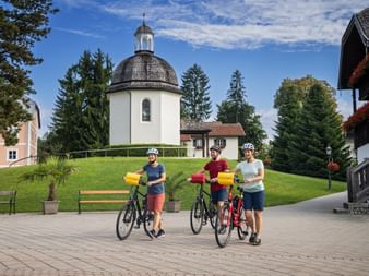 Three cyclists with helmets and bikes stand on a paved square in front of the white Silent Night Chapel with dark dome in Oberndorf.