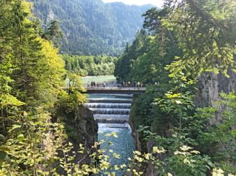 Lechfall waterfall cascading over stepped concrete weir with tourists on viewing platform above, surrounded by lush green forest and mountains.
