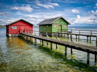 Red and green wooden boathouses on wooden pier extending into Ammersee lake, with clear water, blue sky, and distant sailboats.