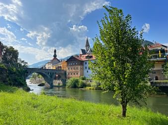 Steinbrücke über Fluss in Murnau mit bunten historischen Gebäuden, Kirchturm und grünem Baum im Vordergrund unter blauem Himmel.