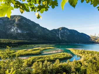 Panoramic view of Sylvensteinsee with turquoise water, winding river delta, and forested mountains in the Bavarian Alps under blue sky.