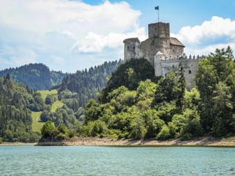 Czorsztyn Castle perched on a forested hill above the turquoise Czorsztyn reservoir, with green mountains in the background under a blue sky.