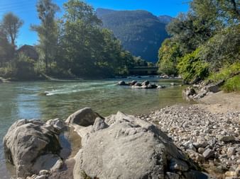 Rocky shore of Saalach River with clear turquoise water flowing past. Forested mountains rise in background under blue sky near Lofer.