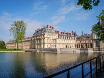 Fontainebleau Palace with cream-colored facade and mansard roof reflected in a large pond. Black railing in foreground, green trees frame the scene.