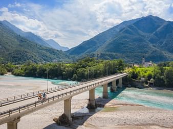Cyclists crossing a concrete bridge over the turquoise Tagliamento River near Venzone, with forested mountains and a small village in the background.