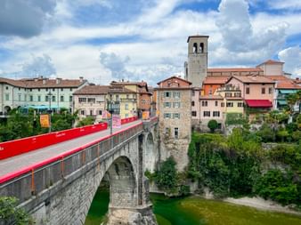 Steinbrücke mit roter Absperrung über grünem Fluss in Cividale. Bunte historische Gebäude und Glockenturm unter bewölktem Himmel.