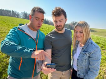 Eurobike-Mitarbeiter in türkisfarbener Jacke zeigt jungem Paar Informationen auf Smartphone in grüner Wiesenlandschaft bei Bozen.