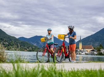 Zwei Radfahrer mit roten Rädern und gelben Taschen pausieren am Tegernsee. Kirche mit gelbem Turm und Berge im Hintergrund sichtbar.