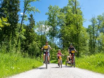 Vierköpfige Familie radelt auf Schotterweg entlang des Drau-Radwegs in Kärnten, umgeben von grünen Bäumen und Gras unter blauem Himmel.