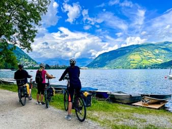 Drei Radfahrer mit Rädern stehen am Zeller See und blicken auf den blauen See mit Bergen und grünen Hängen unter bewölktem Himmel.