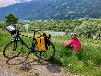 Radfahrer in rosa Jacke sitzt im Gras neben Tourenrad mit gelben Packtaschen und blickt über das Drautal mit Bergen im Hintergrund.