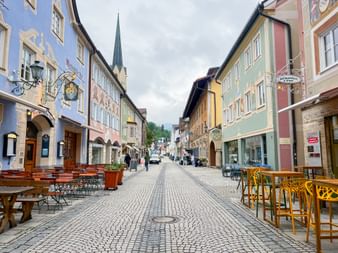 Cobblestone pedestrian street in Garmisch with colorful historic buildings, outdoor seating, and a church spire visible in the background.