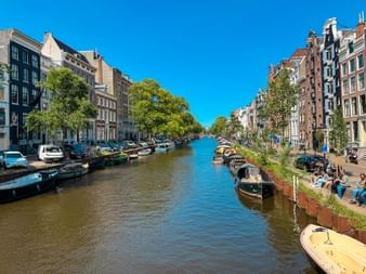 Amsterdamer Gracht gesäumt von traditionellen holländischen Häusern, Booten und Bäumen unter blauem Himmel. Menschen entspannen am Ufer.