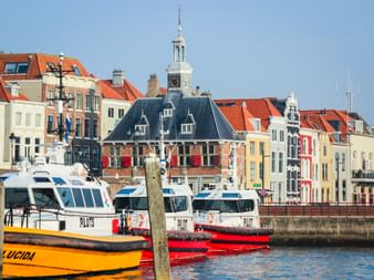 White and yellow pilot boats moored in Vlissingen harbor with colorful Dutch buildings and a historic building with a clock tower in the background.