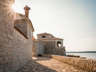 Stone buildings with terracotta roofs along the waterfront in Novigrad, Istria. A cobblestone path leads past historic walls toward the sea.