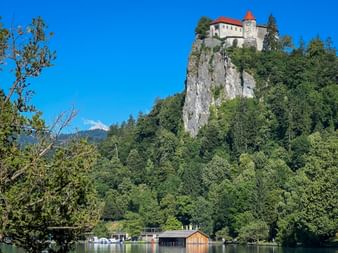 Burg Bled thront auf einer steilen Felsklippe über dem Bleder See, umgeben von dichtem grünen Wald. Bootshäuser am Ufer sichtbar.