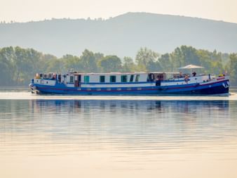 Blue and white river cruise ship sailing on calm water in Provence-Camargue with green trees and hills in the background.