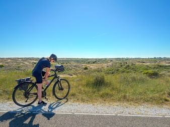Radfahrer in schwarzer Kleidung mit Tourenrad auf Küstenweg bei Noordwijk. Dünenlandschaft mit Gras und Sträuchern unter blauem Himmel.