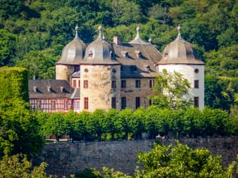 Schloss in Gemünden mit vier runden Türmen und Kuppeldächern, umgeben von Steinmauern und grünen Bäumen an einem bewaldeten Hang.
