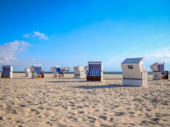 Traditional German beach chairs scattered on sandy beach at Sylt. Blue and white striped wicker baskets numbered 104 visible under clear sky.